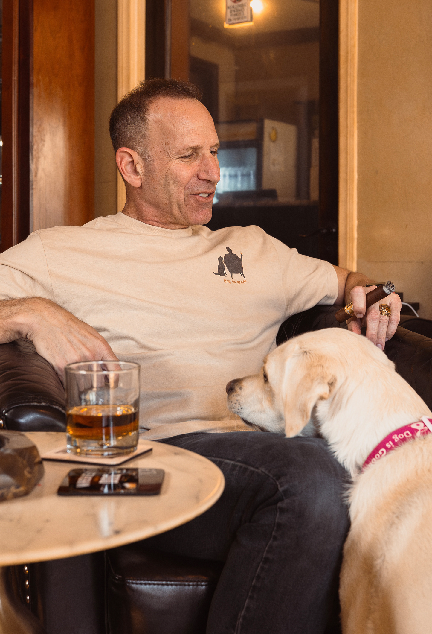 Man wearing tan Conversation Not Required T-shirt with dog silhouette, sitting with whiskey and dog nearby