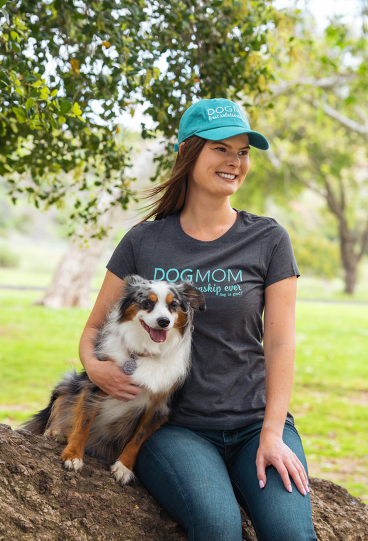 Woman wearing turquoise Dog Mom Hat and matching Dog Mom T-shirt holding a fluffy dog outdoors
