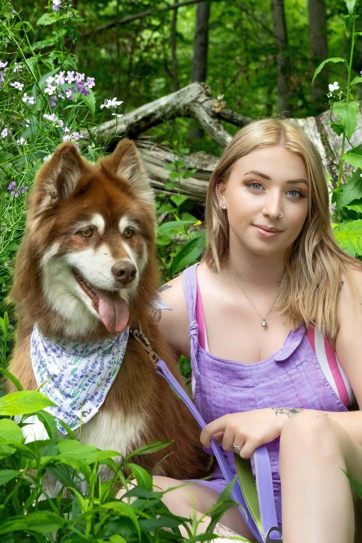Dog wearing lavender blooms dog bandana sitting next to woman in lavender dress in a green outdoor setting