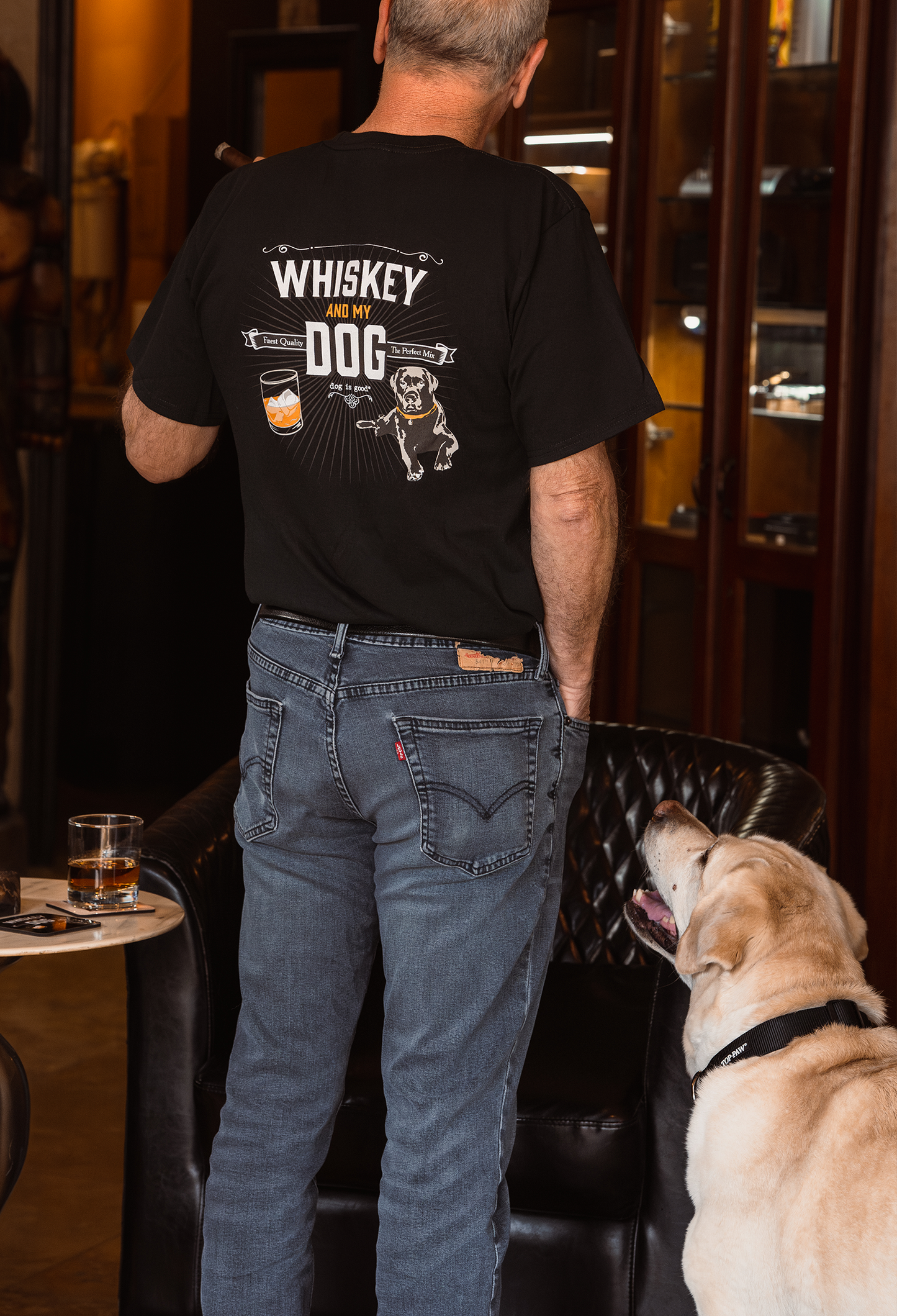 Man wearing black "Whiskey and My Dog" T-shirt with a dog looking up inside a cozy room