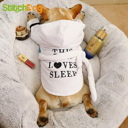 Small dog wearing white hooded bath robe with "This Loves Sleep" text lying on soft blanket