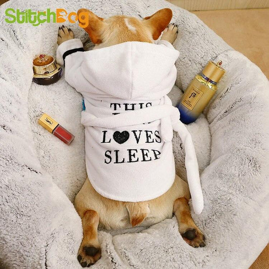 Small dog wearing white hooded bath robe with "This Loves Sleep" text lying on soft blanket