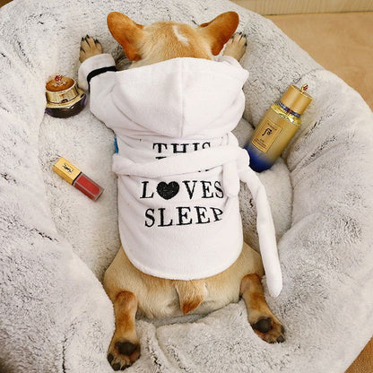 Small dog wearing white bath robe with hood lying on soft bed with beauty products around