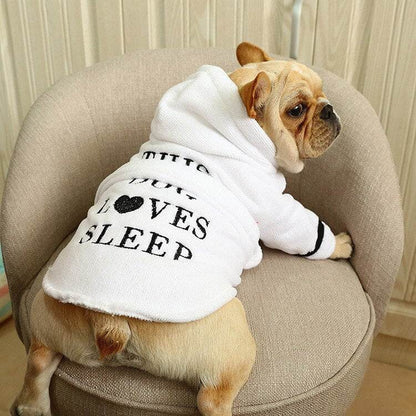 Small dog wearing white hooded bath robe with "Love Sleep" text sitting on beige chair