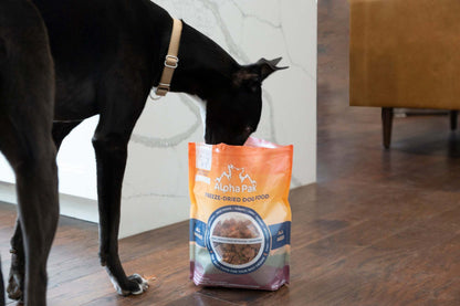 Black dog sniffing a bag of Alpha Pak Premium Raw Freeze Dried Dog Food on wooden floor