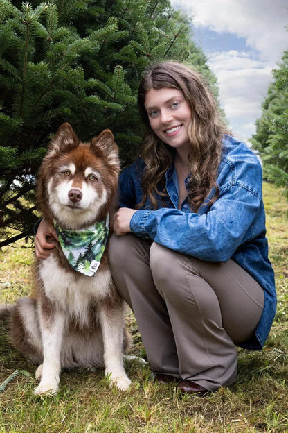 Dog wearing Evergreen Explorer Dog Bandana with green pine forest pattern sitting next to smiling woman outdoors