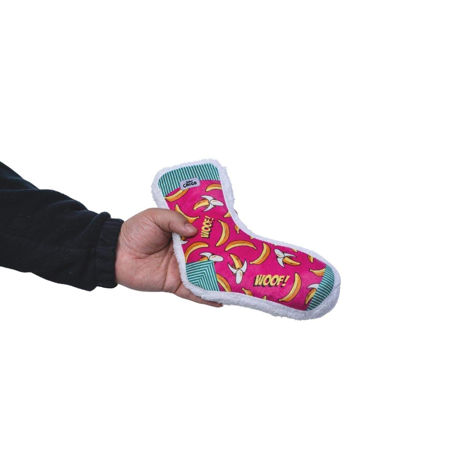 Hand holding pink Squeaky Sock Plush Dog Toy with banana pattern and "WOOF!" text designed for moderate chewers