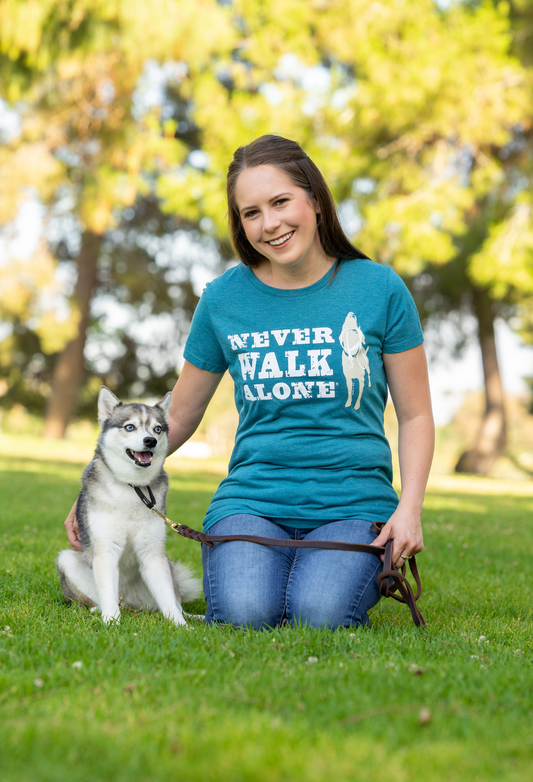Woman wearing teal Never Walk Alone t-shirt sitting on grass with her dog in a park