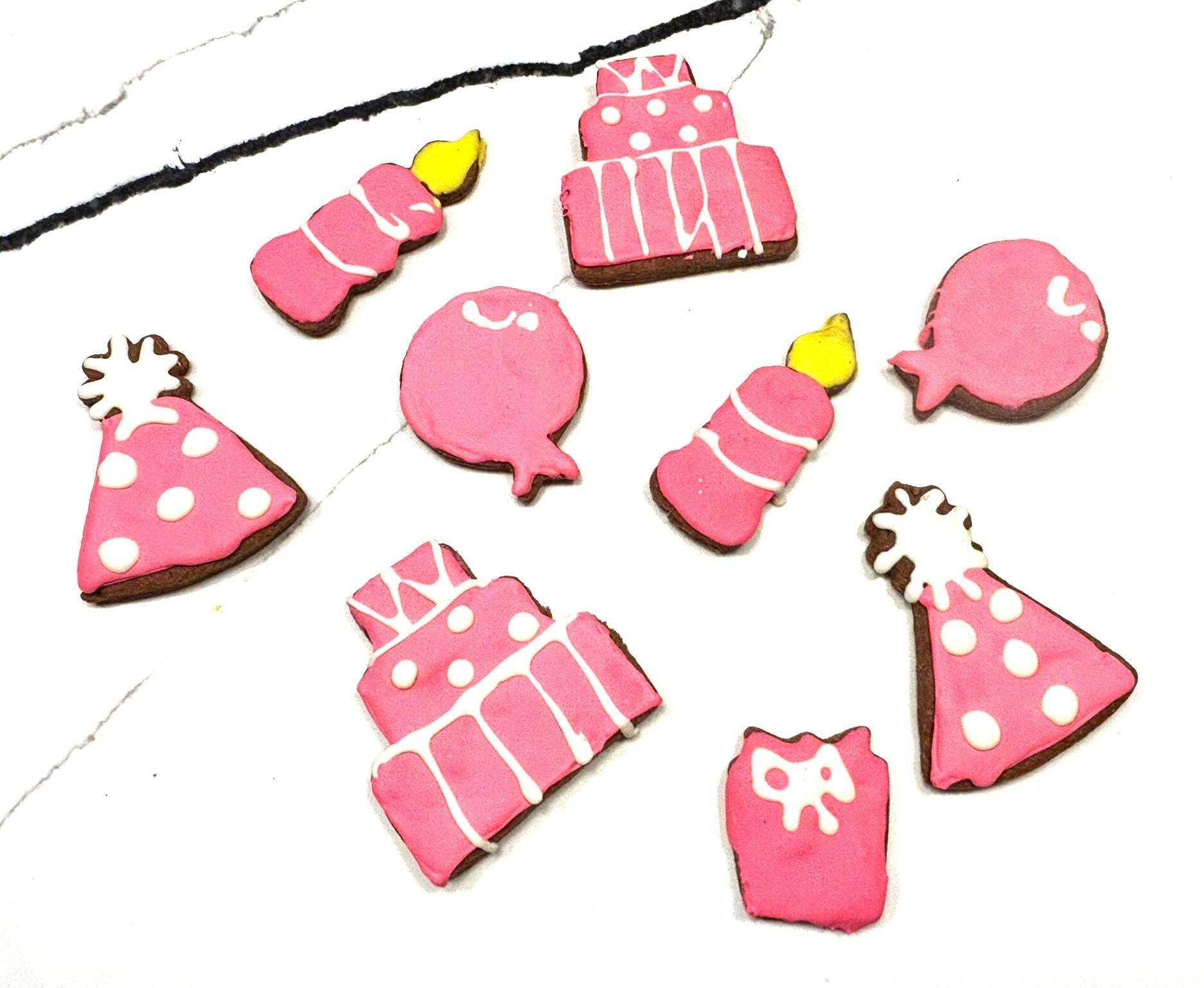 Pink birthday girl dog cookies shaped like party hats, candles, balloons, and cakes with white detailing on white marble background