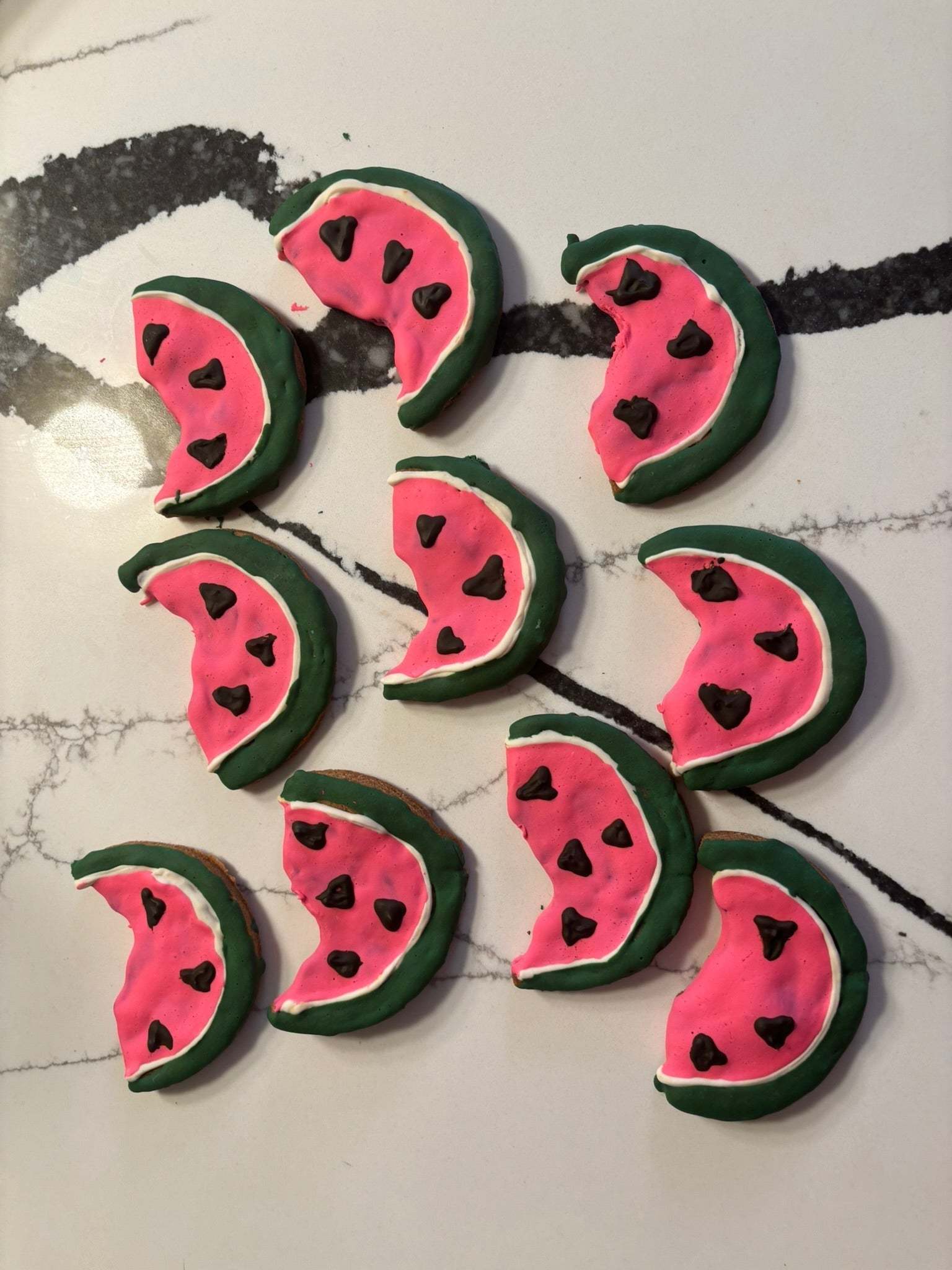 Organic Watermelon Dog Cookies shaped like watermelon slices with pink, green, and black colors, laid out on a marble surface.