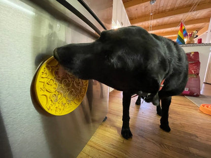Black dog licking yellow Rubber Duckies emat lick mat attached to stainless steel appliance with suction cups in kitchen