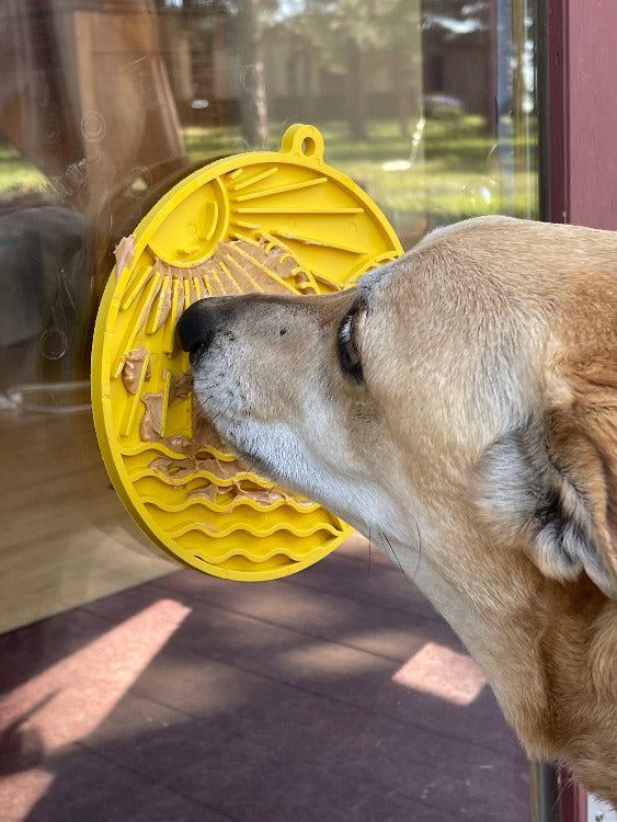 Dog licking yellow beach-themed Sun n' Sea eMat lick mat with suction cups on glass door for enrichment and anxiety relief