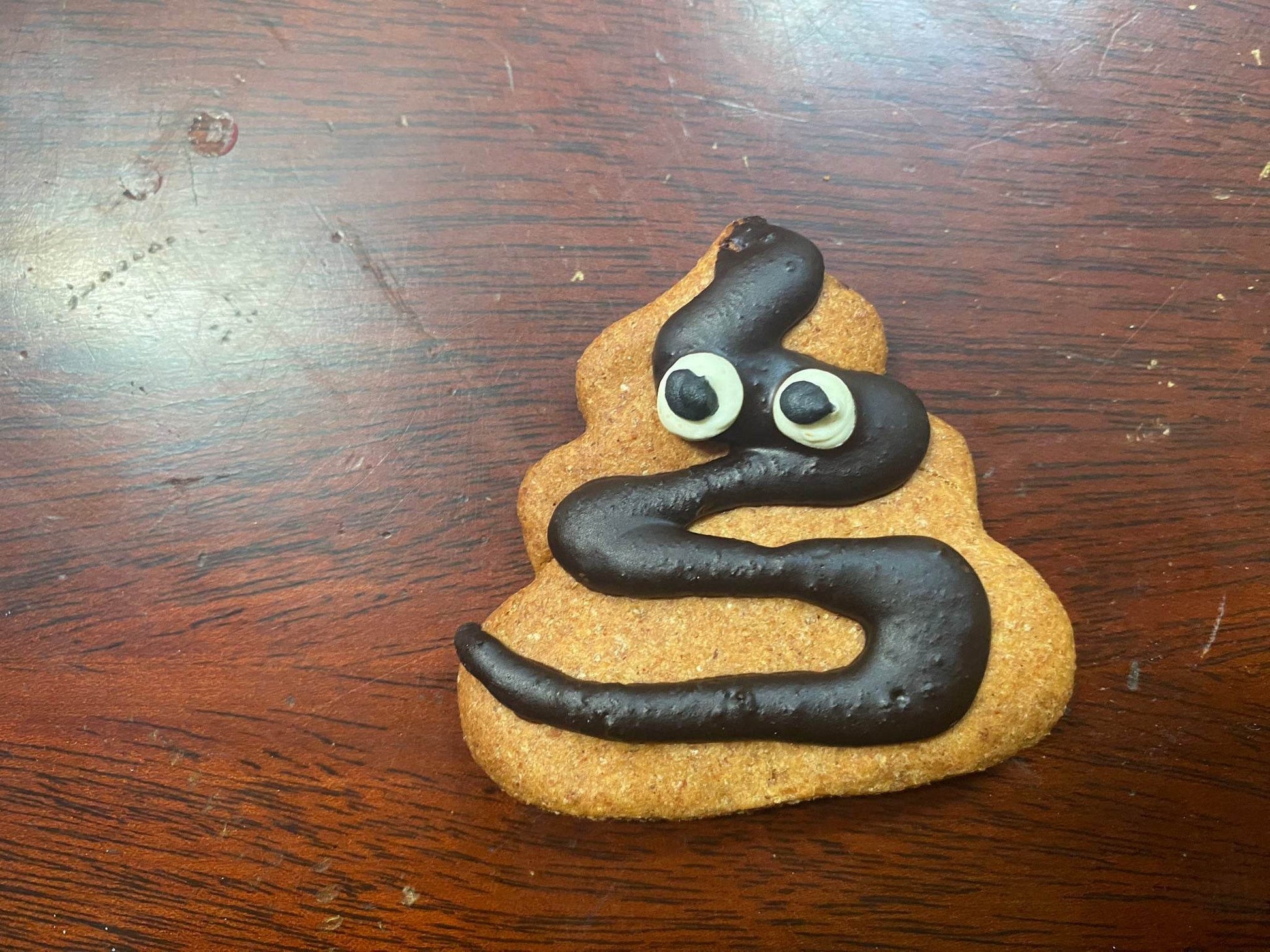 Poop Organic Dog Cookie shaped like poop emoji with chocolate icing eyes and swirl on wooden surface
