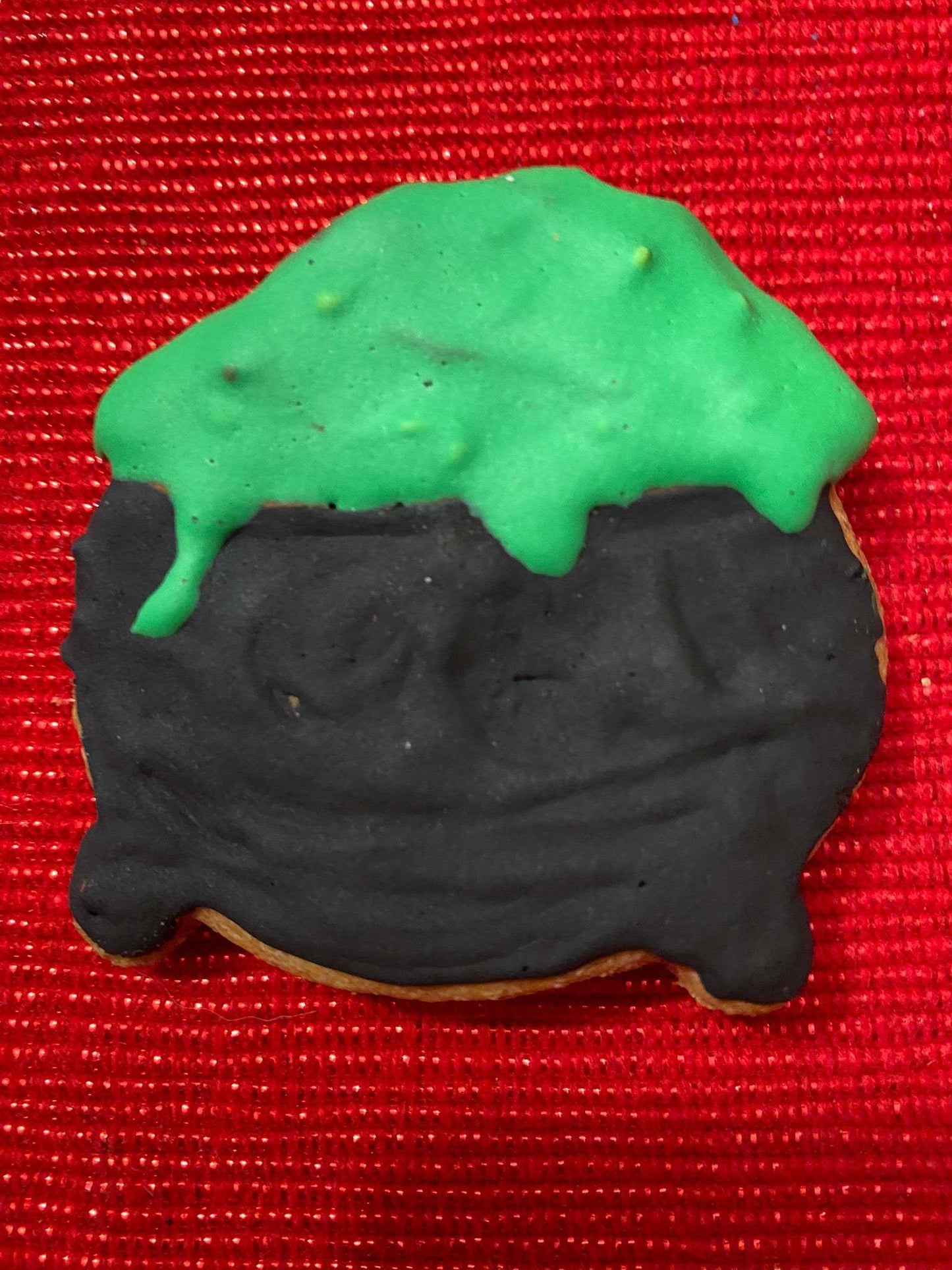 Halloween-themed organic dog cookie shaped like a bubbling witch's cauldron with black and green icing on a red textured background