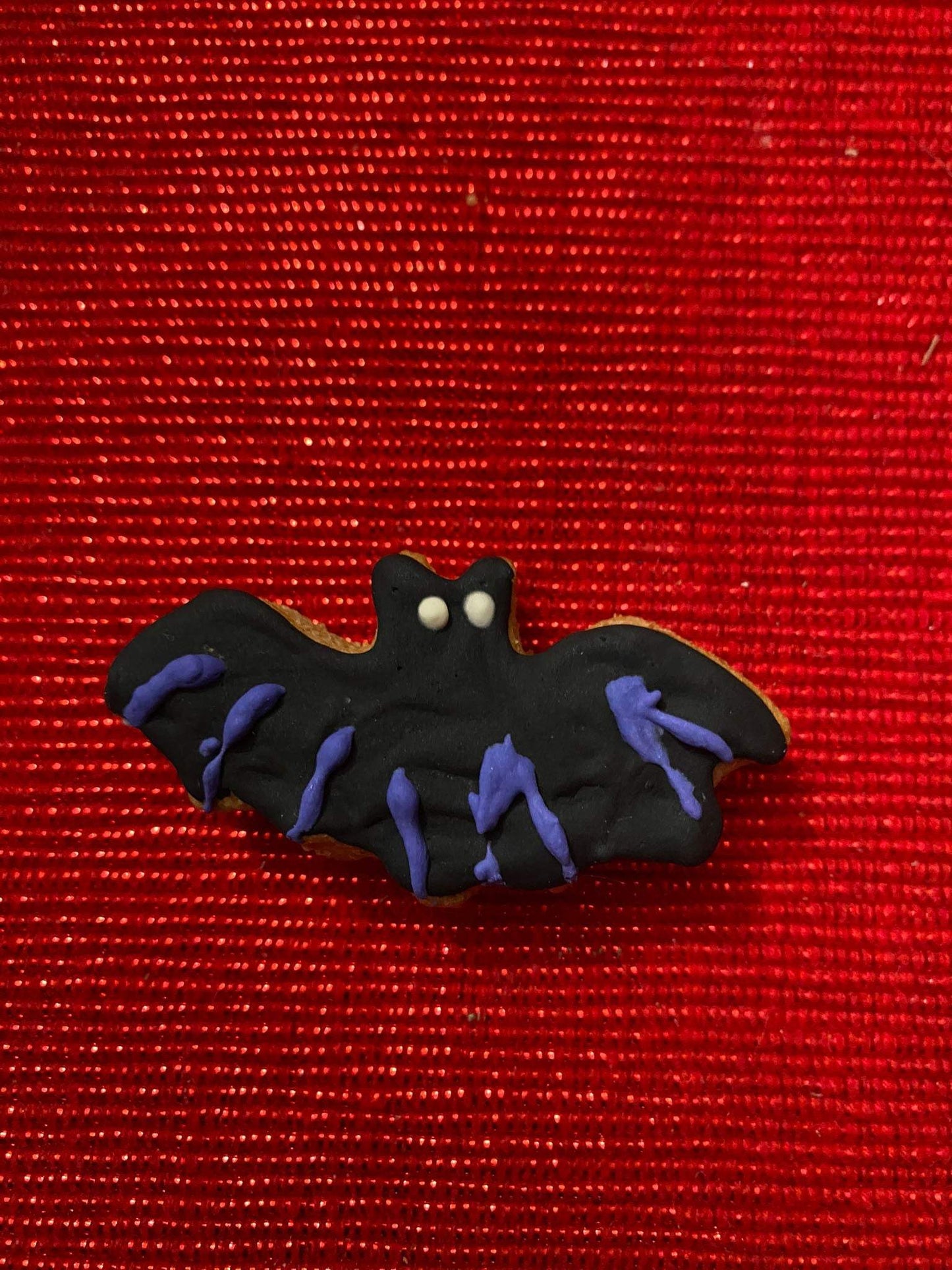 Halloween organic dog cookie shaped like a black bat with purple icing on red textured background