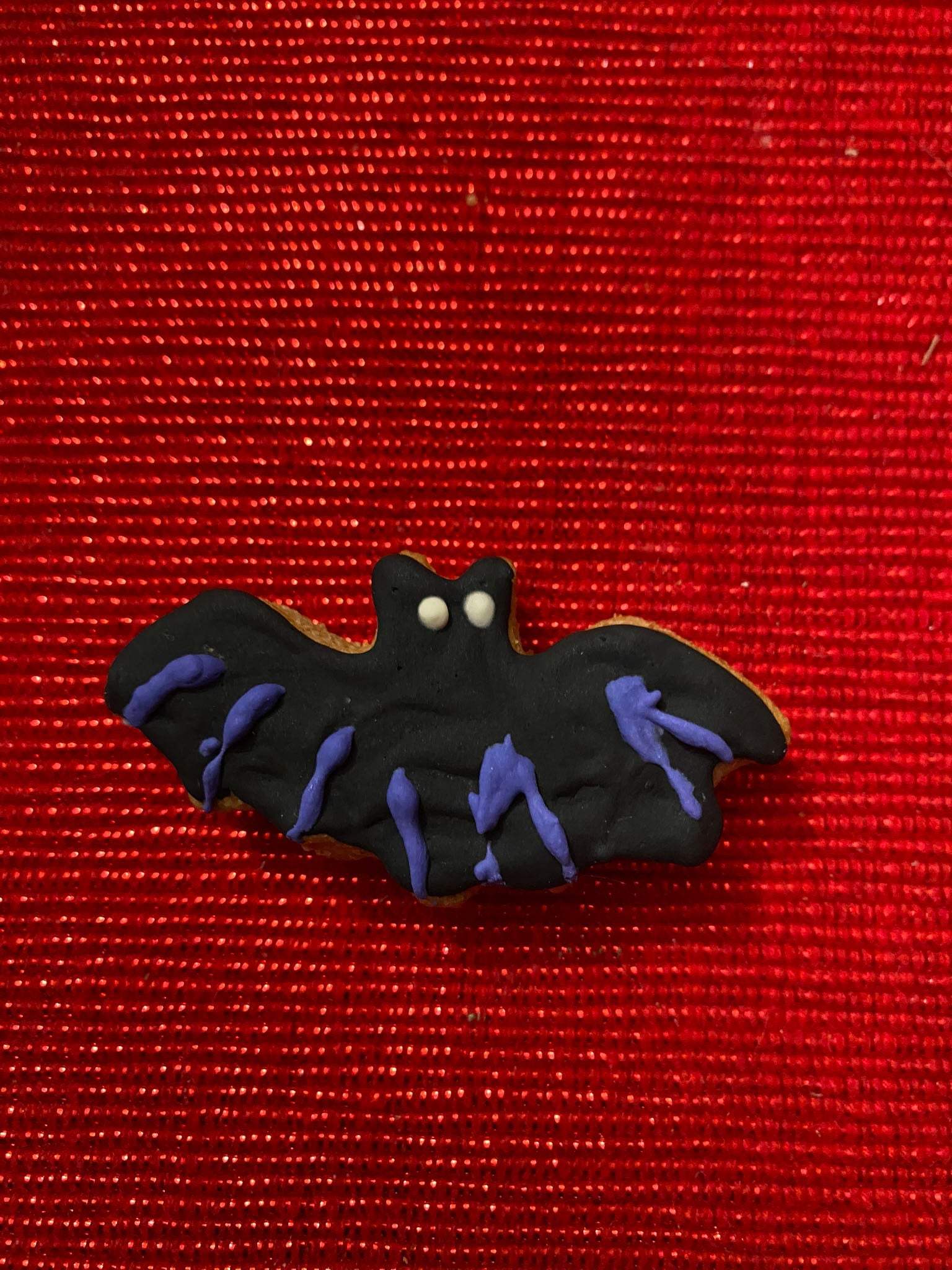 Halloween organic dog cookie shaped like a black bat with purple icing on red textured background