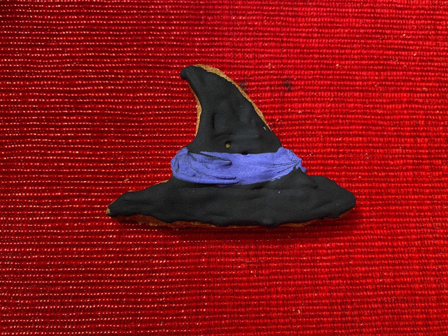 Black witch hat shaped organic Halloween dog cookie with purple icing band on red textured background
