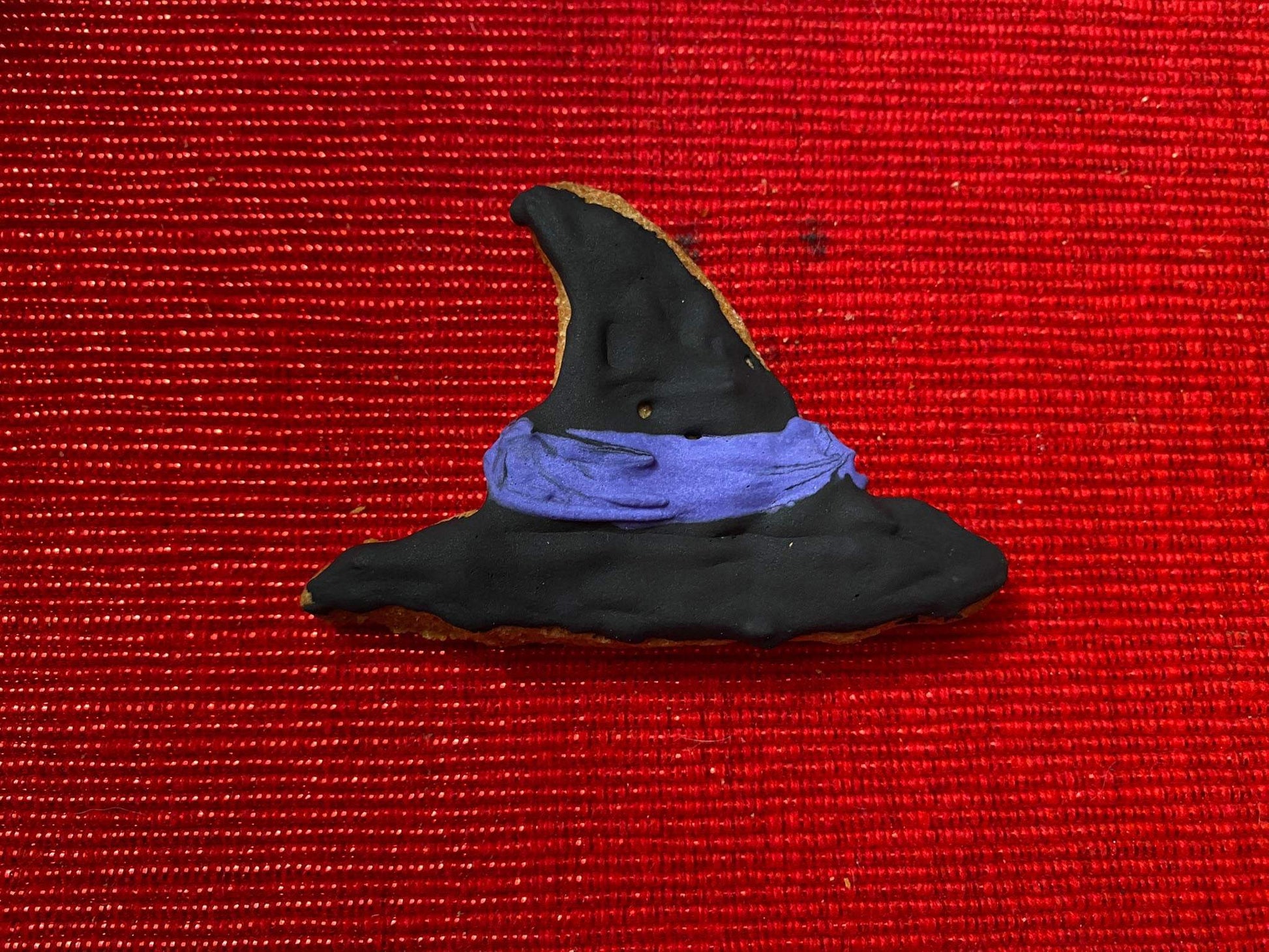 Black witch hat shaped organic Halloween dog cookie with purple icing band on red textured background