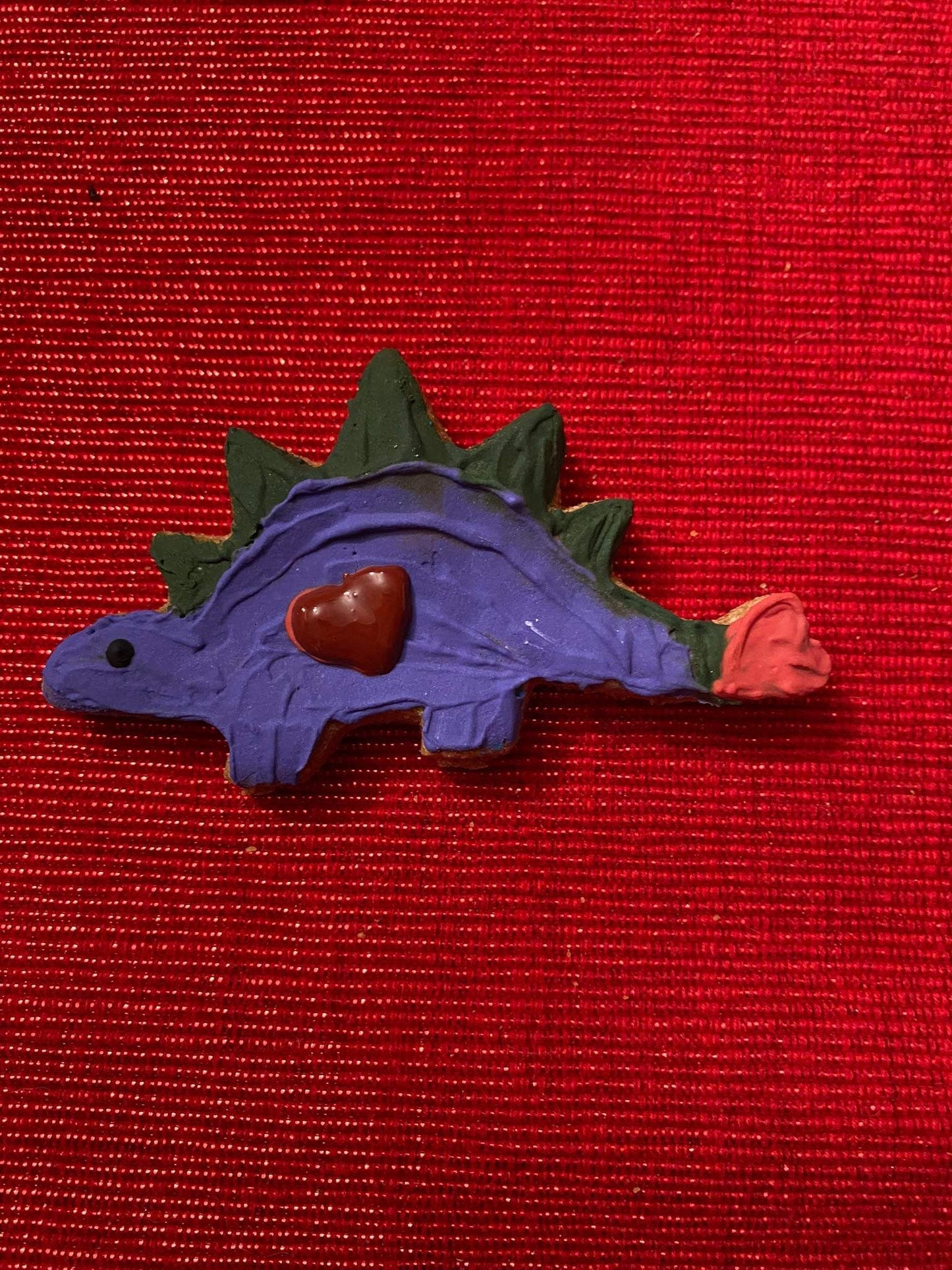 Purple dinosaur-shaped organic dog cookie with a heart decoration on red textured background