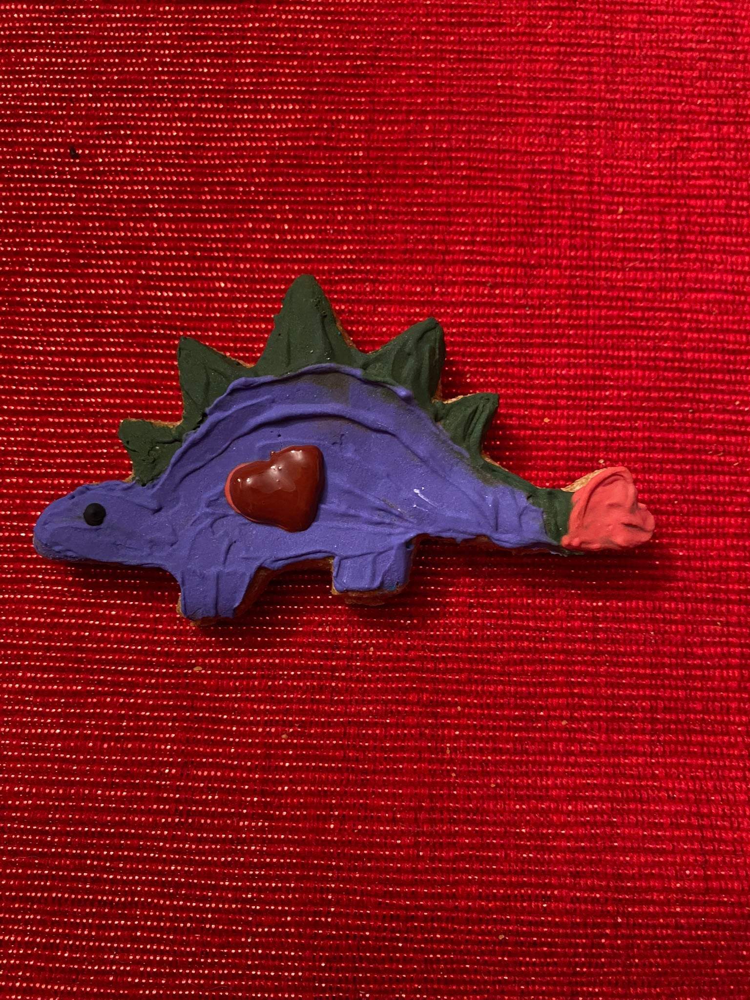 Purple dinosaur-shaped organic dog cookie with a heart decoration on red textured background