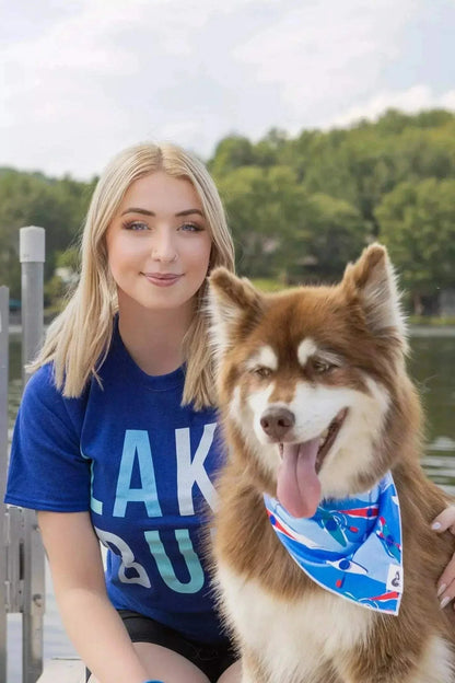 Dog wearing a vibrant blue K9 Kayak Dog Bandana with kayak pattern sitting next to a woman by the lake