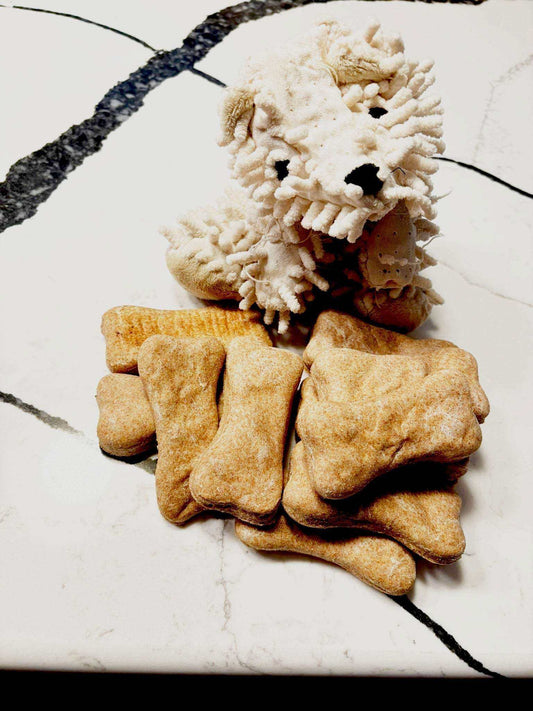 Cheddar Paw Bones bone-shaped dog treats in golden brown with a plush white dog toy on white marble surface