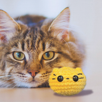 Cat resting head beside small yellow weighted cat toy Pippa the Chonk with plush round shape and black stitched face.
