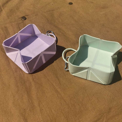 Two compact foldable pet food and water bowls in pastel purple and green with attached carabiners on brown surface